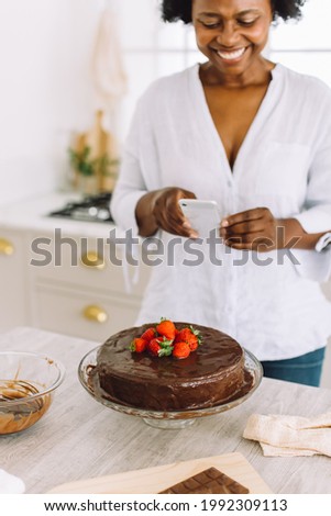 Mature woman taking pictures of a cake in the kitchen. Female capturing photos of chocolate cake with her mobile phone.