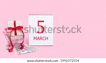 5th day of march. A gift box in a shopping trolley, dollars and a calendar with the date of 5 march on a pink background. Spring month, day of the year concept.