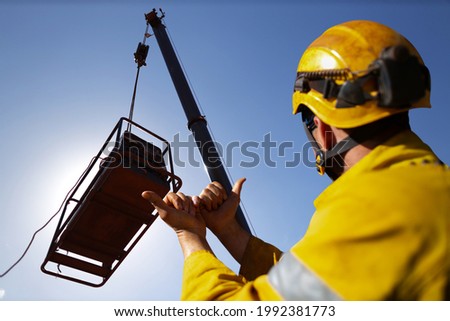 Safe work practice rigger wearing fall protection helmets giving crane operator hand signal by holding up two hand against each other pointing boths thump out meant crane boom is giging out slowly    