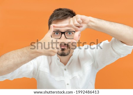 Portrait of attentive curious bearded happy male in white shirt looking at camera, focusing through frame made of fingers. Indoor studio shot isolated on orange background. 