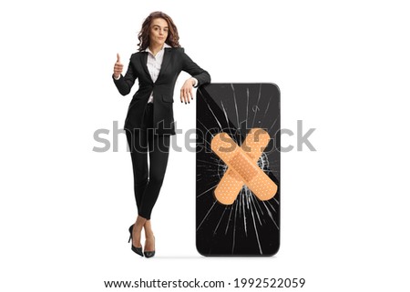 Full length portrait of a young businesswoman with a thumb up gesture leaning on a cracked smartphone isolated on white background