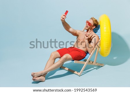 Full length young man in red shorts swimsuit glasses sit in chair inflatable ring do selfie shot mobile cell phone show v-sign isolated on pastel blue background Summer vacation sea sun tan concept