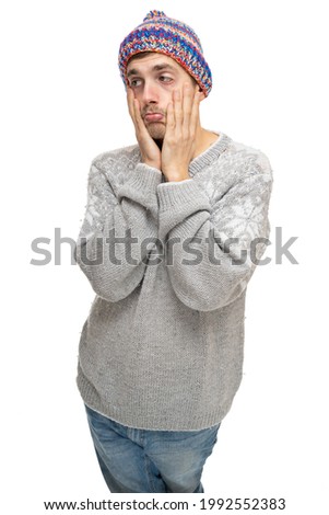 Young handsome tall slim white man with brown hair pulling his face in gray jumper with colorful winter hat isolated on white background