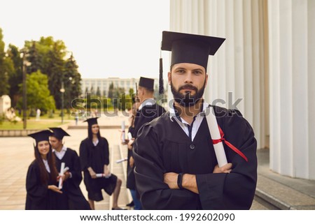 Portrait of a confident and joyful student with a beard in the uniform of a university graduate and with a diploma in hand. Guy poses with crossed arms on the background of classmates.