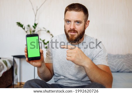 European bearded man with red hair and big blue eyes shows off something on smartphone with green screen to camera.