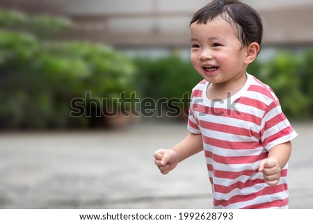 Portrait of an asian boy (toddler) running toward and smiling with happy and fun face while playing outdoor. A Child wear striped shirt in red and white color. Head and hair is wet by sweat. Royalty-Free Stock Photo #1992628793