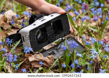 Hand holding 3D Virtual reality headset glasses with front of lens opened in spring blue and purple wild flowers. VR tech gear goggles close-up