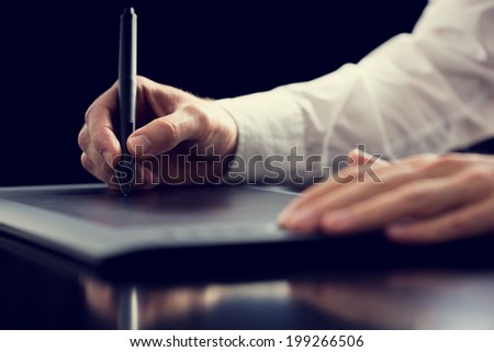 Retro effect faded and toned image of a graphic designer working with digital tablet pen, over black background.