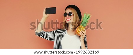 Summer portrait of happy smiling young woman with pineapple taking a selfie picture by smartphone on a brown background