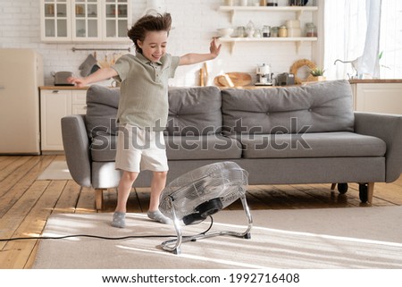 Little boy play with strong wind blow from ventilator or industrial fan at home in cozy living room. Small kid alone with retro air conditioner enjoy refreshing fresh breeze. Child has fun with cooler Royalty-Free Stock Photo #1992716408