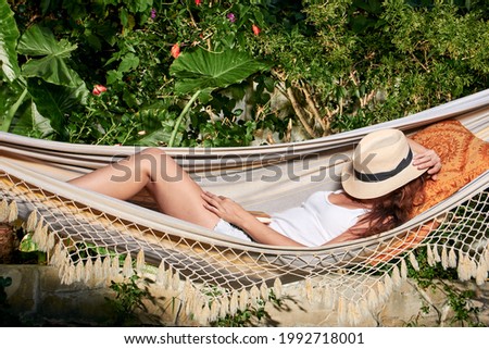 Woman lying suntanning on a hammock wearing a trendy hat covering her face from the sun in a tropical garden in a relaxation or vacation concept