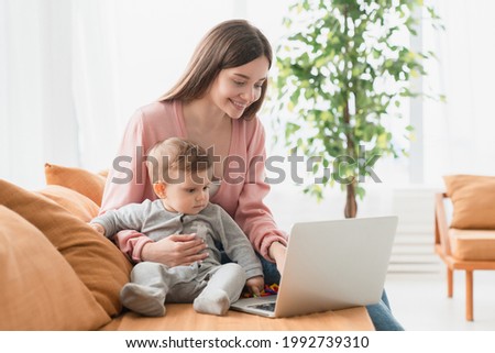 Young single mother mom with toddler newborn baby infant son using laptop, searching web, watching cartoons together. Working from home on maternity leave, freelancer, looking for kids goods