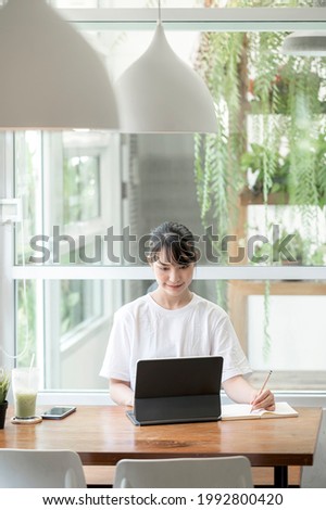 Portrait of young beautiful woman working on tablet computer and writing on notebook while sitting at the table, online learning concept.