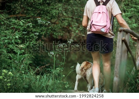 a young brunette with pink backpack on her back stands on a bridge by the river. a white-and-red dog runs around nearby. Rear view.