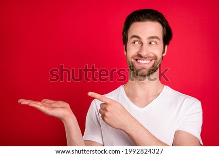 Photo of joyful young brown hairdo man hold point look empty space wear white t-shirt isolated on red color background