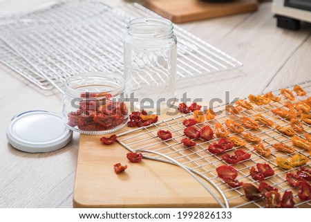 Dehydrate dried yellow and red tomatoes on stainless steel dehydrator tray and in glass jar for long term pantry in domestic kitchen on light wooden background