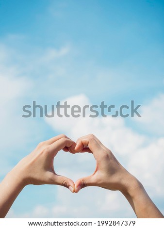Heart shape hand on blue sky and fluffy cloud background, concept of love, relationship and romantic, vertical style. Female hand making finger love sign with copy space.