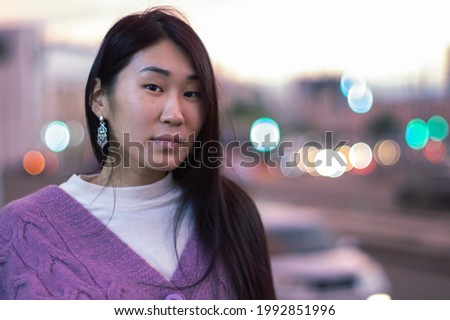 portrait of long haired asian woman in vintage sweater against blurry city lights background during sunset. close-up