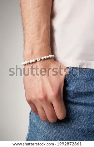   Cropped close-up shot of male wrist with beaded bracelet made of white marble stone and decorated with silver beads and charm with letter G. Man in beige t-shirt put his hand in the pocket.         