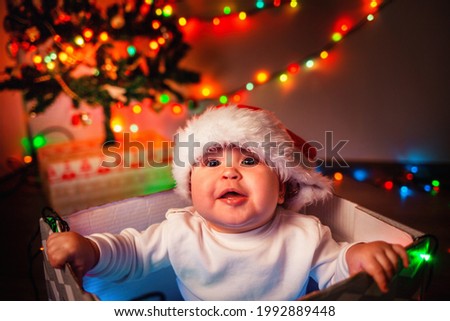 Christmas. Portrait of a cute baby in a Christmas hat sitting in a gift box. A garland lights up in the background. The concept of winter Christmas holidays.