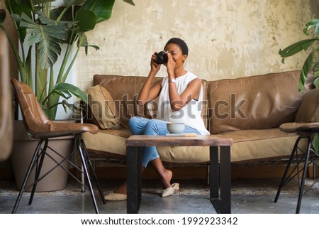 Shot of a young woman taking pictures on a vintage camera. She is sitting on the sofa in the room with earthy colors.
