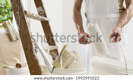 hands of house painter man decorator work of home to renovate, using roller paint and holding white bucket, a wooden ladder with paint brushes as background, close-up Royalty-Free Stock Photo #1992991022