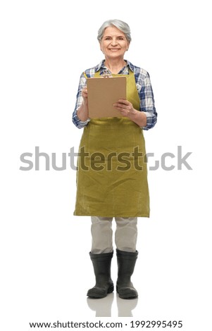 gardening, farming and old people concept - portrait of smiling senior woman in green garden apron and rubber boots with clipboard over white background