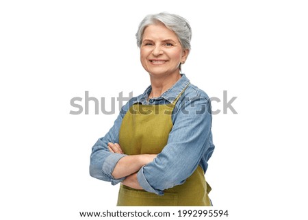 gardening, farming and old people concept - portrait of smiling senior woman in green garden with apron crossed arms over white background