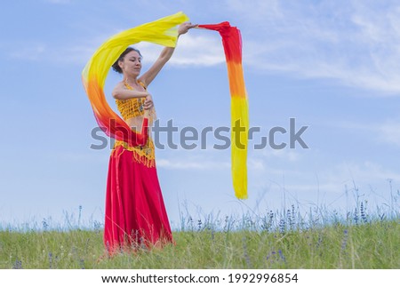 A young happy woman in national costume with fiery fans in her hands is dancing an incendiary oriental dance on a green lawn against a blue sky 