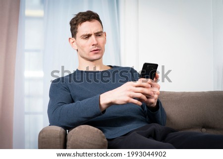 Portrait of sad unpleasant unhappy guy, young frowning man looking at screen of his cell mobile phone, using smartphone with negative face, sitting at home in living room on sofa or couch. Bad news Royalty-Free Stock Photo #1993044902
