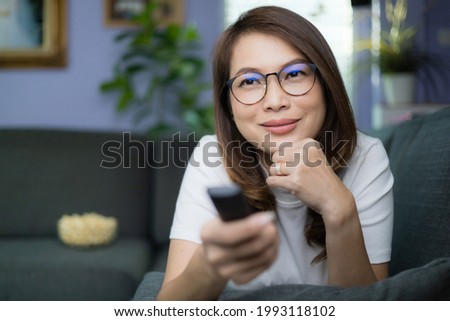 Cute Asian woman laying on sofa with relax and cozy gesture holding remote control aiming to tv, watching television and smile with fun and happy with popcorn blur in background. Hobby and relax idea.