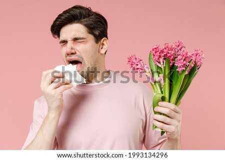 Sick allergic man has red eyes runny stuffy sore nose suffer from pollen allergy symptoms hay fever hold bloom flower plant napkin reaction on trigger isolated on pastel pink color background studio Royalty-Free Stock Photo #1993134296