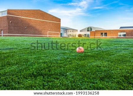 Old and deflate orange football on green turf with sunlight at school building background, depth of field. Outdoor recreation space for students. Landscape view of empty soccer field and unused ball.