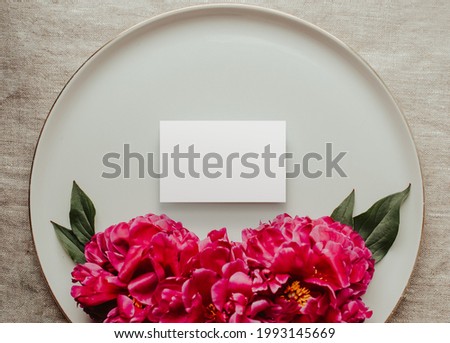 Blooming branches with peony flowers and buds. Blank business card mockup on ceramic plate. Feminine wedding and red flowers of peonies. Gray linen tablecloth fabric background. Botanical and luxury