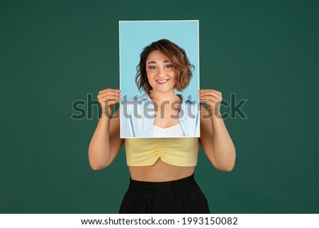Different emotions, mood. One young beautiful woman holding her portraits pictures isolated on green background. Concept of human emotion, facial expression, show, youth. Copy space for ad