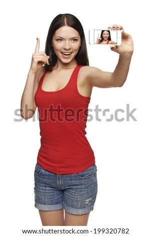 Happy excited young winking girl taking pictures of herself through cell phone, over white background