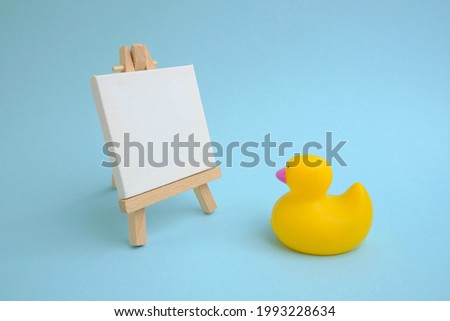 A yellow rubber little duck looks at a molbert with a white canvas.