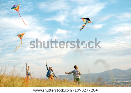 Smiling girls and brother boy with flying colorful kites - popular outdoor toy on the high grass mountain meadow. Happy childhood moments or outdoor time spending concept image. Royalty-Free Stock Photo #1993277204