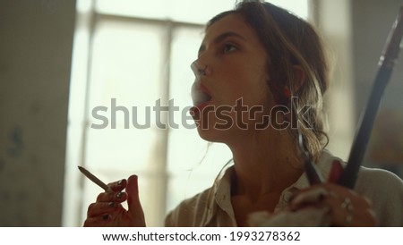 Talented young woman painter smoking in studio. Creative girl blowing smoke out indoors. Female artist holding brush at workplace on light window background.