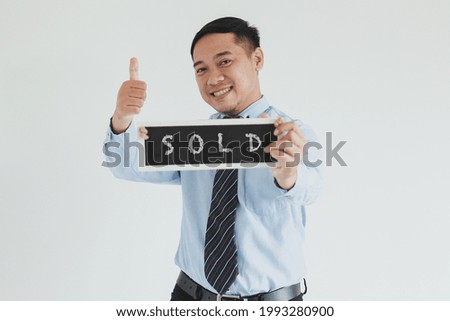 Smiling sales man wearing blue shirt and tie posing with thumb up and showing "SOLD" sign board at camera on white background