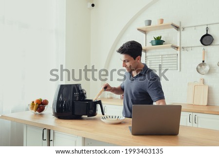 A portrait of a Hispanic man  learning the air fryer machine manual on the Internet in home. Adult men looking for recipes on laptop at modern kitchen.  He learning to cook with online course Royalty-Free Stock Photo #1993403135