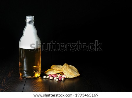glass bottle with light beer with large foam, with salty chips and nuts on a dark wooden table on a black background with beautiful lighting and reflection on the table