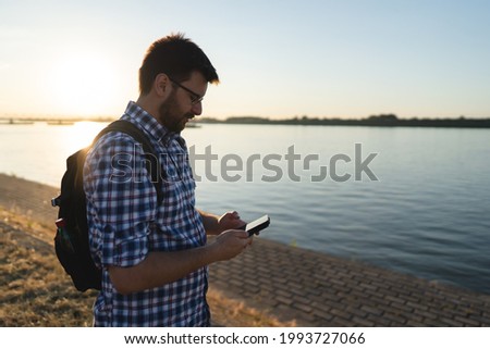 Side view waist up portrait of adult caucasian man tourist using mobile phone while standing by the Danube river in sunset in sunny summer day wearing shirt and backpack real people copy space