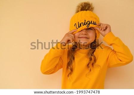 A cute little girl pulls on an orange cap with the words "unique" over her eyes and grimaces against. Copy space. A unique child. Children of the new generation. Fashionably dressed child