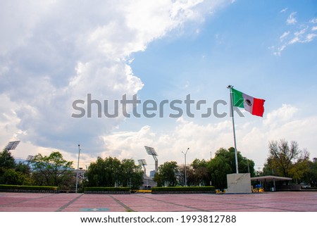 Wavy flag from Mexico in empty public square with trees and slightly cloudy sky as background. Green, white and red flag waving with the wind under bright skyline. Mexican patriotic symbols