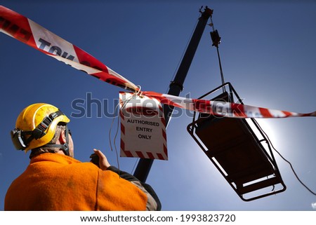 Safety work practice red and white warning danger tag tape sign applying on exclusion dropped zone where rigger operating 2 way radio with crane operator during lifting heavy load at open field  Royalty-Free Stock Photo #1993823720