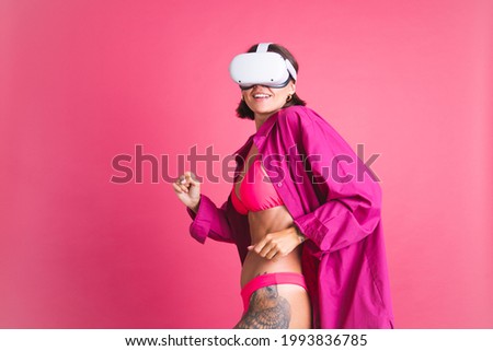 Beautiful fit tanned sporty woman in bikini and shirt in virtual reality glasses on pink background