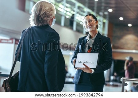 Private driver wearing face shield standing at airport terminal holding a  signboard and receiving a traveler. Female chauffeur with a sign at airport arrival gate.