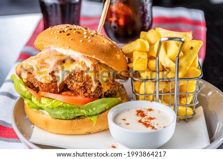 A chicken parma burger with lettuce and tomato on a white plate with french fries in a wire basket and dipping sauce