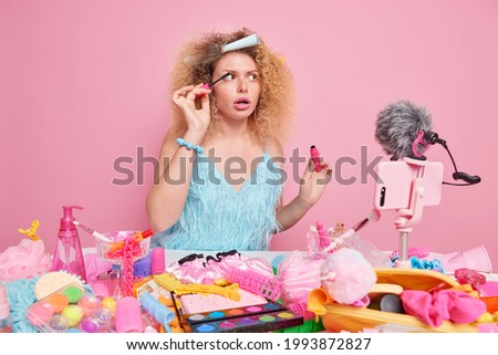 Curly haired female model applies mascara gives professional advice about doing makeup wears blue dress surrounded by beauty products records video for social media blog isolated on pink wall